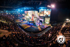 Zu sehen ist eine dunkle Halle, die mit Zuschauerrängen ausgestattet ist. Auf den Rängen sitzen Menschen, die auf eine Bühne schauen, die am Ende der Halle aufgebaut ist. Auf der Bühne spielen zwei E-Sport-Teams gerade ein Spiel gegeneinander, das auf einem großen Bildschirm in der Mitte über der Bühne auf einer riesigen Leinwand angezeigt wird.