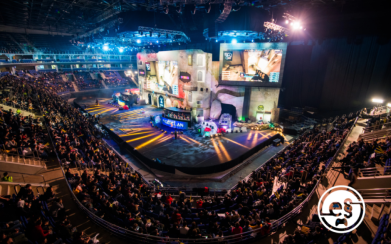 Zu sehen ist eine dunkle Halle, die mit Zuschauerrängen ausgestattet ist. Auf den Rängen sitzen Menschen, die auf eine Bühne schauen, die am Ende der Halle aufgebaut ist. Auf der Bühne spielen zwei E-Sport-Teams gerade ein Spiel gegeneinander, das auf einem großen Bildschirm in der Mitte über der Bühne auf einer riesigen Leinwand angezeigt wird.