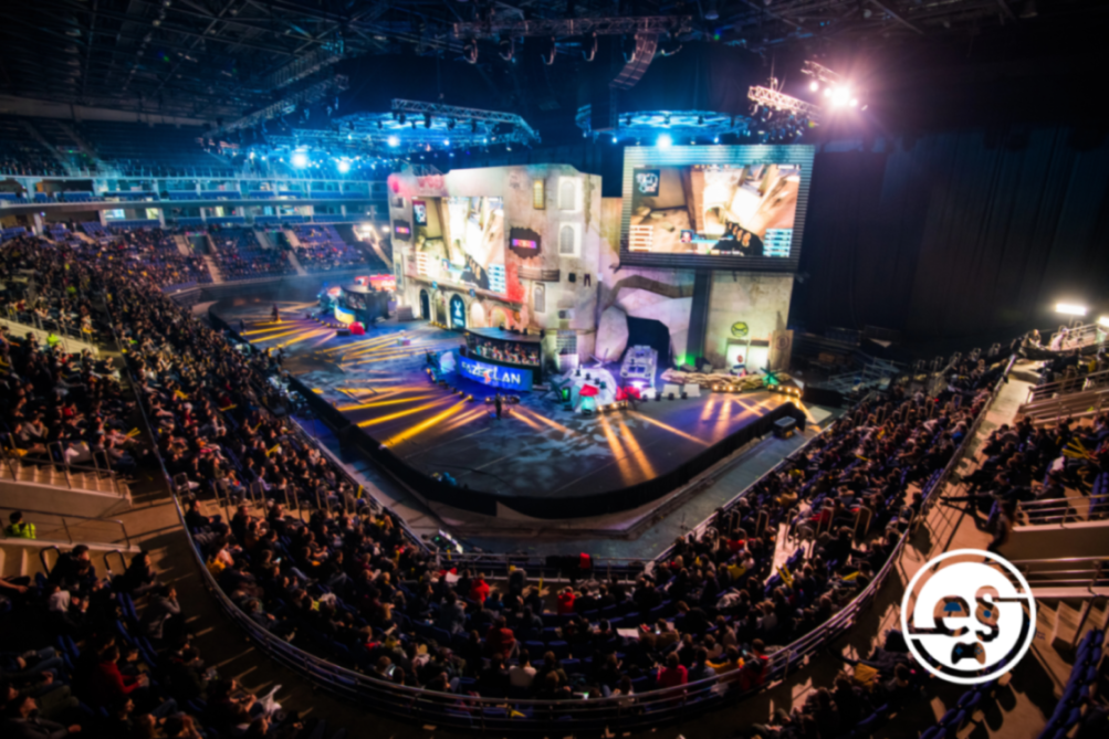 Zu sehen ist eine dunkle Halle, die mit Zuschauerrängen ausgestattet ist. Auf den Rängen sitzen Menschen, die auf eine Bühne schauen, die am Ende der Halle aufgebaut ist. Auf der Bühne spielen zwei E-Sport-Teams gerade ein Spiel gegeneinander, das auf einem großen Bildschirm in der Mitte über der Bühne auf einer riesigen Leinwand angezeigt wird.